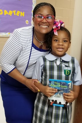 A teacher standing next to a young student holding a book called "The Aquarium"