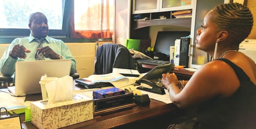 Two educators speak to each other. One is behind a desk with a computer, the other is sitting at the other side of the desk.