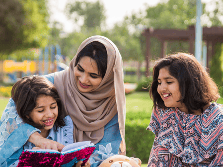 Photo of a mother reading to her two daughters in a park. 