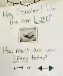 A picture of a poster hanging on a wall that says, "Hey Scholar! Do you even lift? How much are you lifting today?" And then a drawn picture of a barbell with a little weight next to one with a lot of weights. There's a picture of a human brain in the middle after "Do you even lift?"